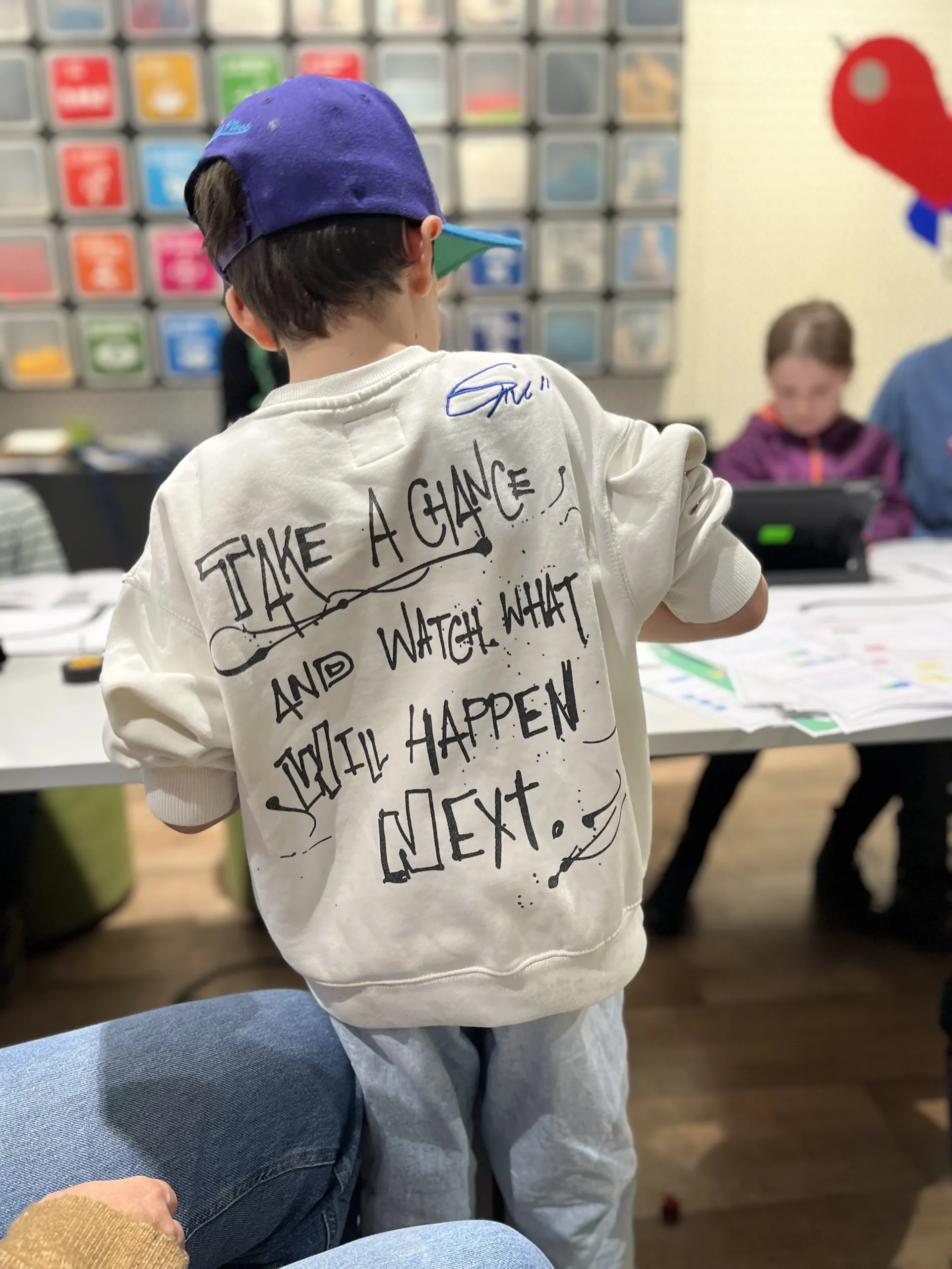 Ein Kind mit blauer Kappe und einem weißen Sweatshirt, auf dem der Spruch 'TAKE A CHANCE AND WATCH WHAT WILL HAPPEN NEXT' steht, in einem Workshop.