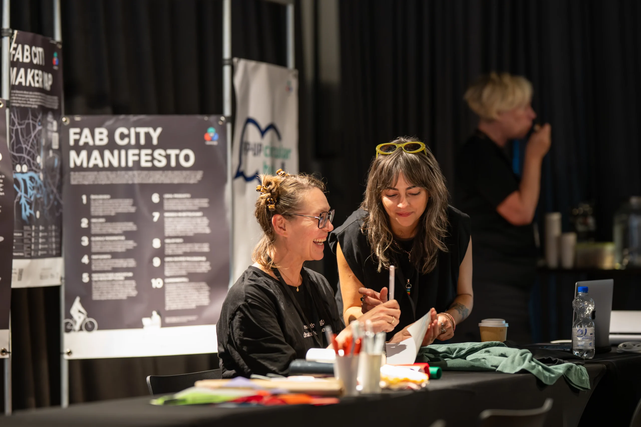 Zwei Frauen interagieren an einem Tisch mit Materialien vor Postern des Fab City Manifestos und der Maker Map.