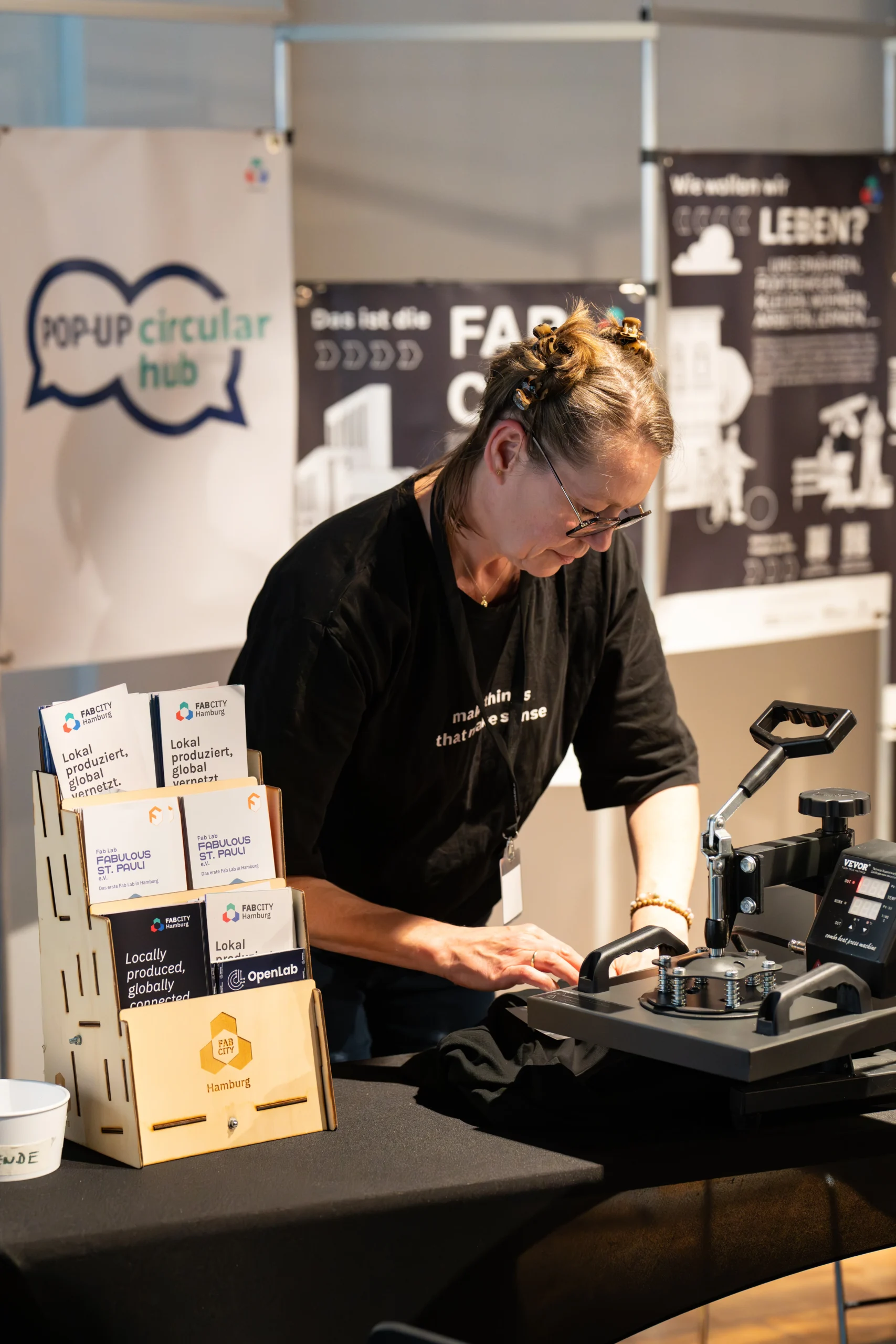 Eine Person bedruckt ein T-Shirt an einer Transferpresse, umgeben von Informationsmaterialien von Fab City Hamburg und dem Pop-Up Circular Hub.