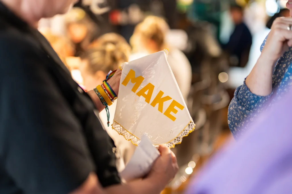 Eine Hand hält ein weißes Tuch mit dem gelben Schriftzug "MAKE", am Handgelenk sind bunte Armbänder zu sehen.