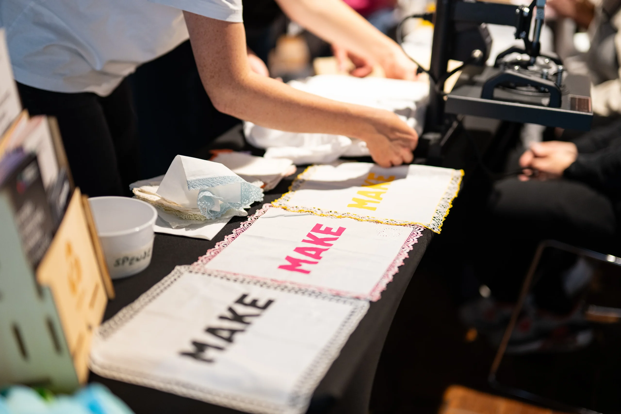 Nahaufnahme eines Tisches mit weißen Tüchern, auf denen das Wort „MAKE“ in verschiedenen Farben zu sehen ist, während Hände im Hintergrund arbeiten.