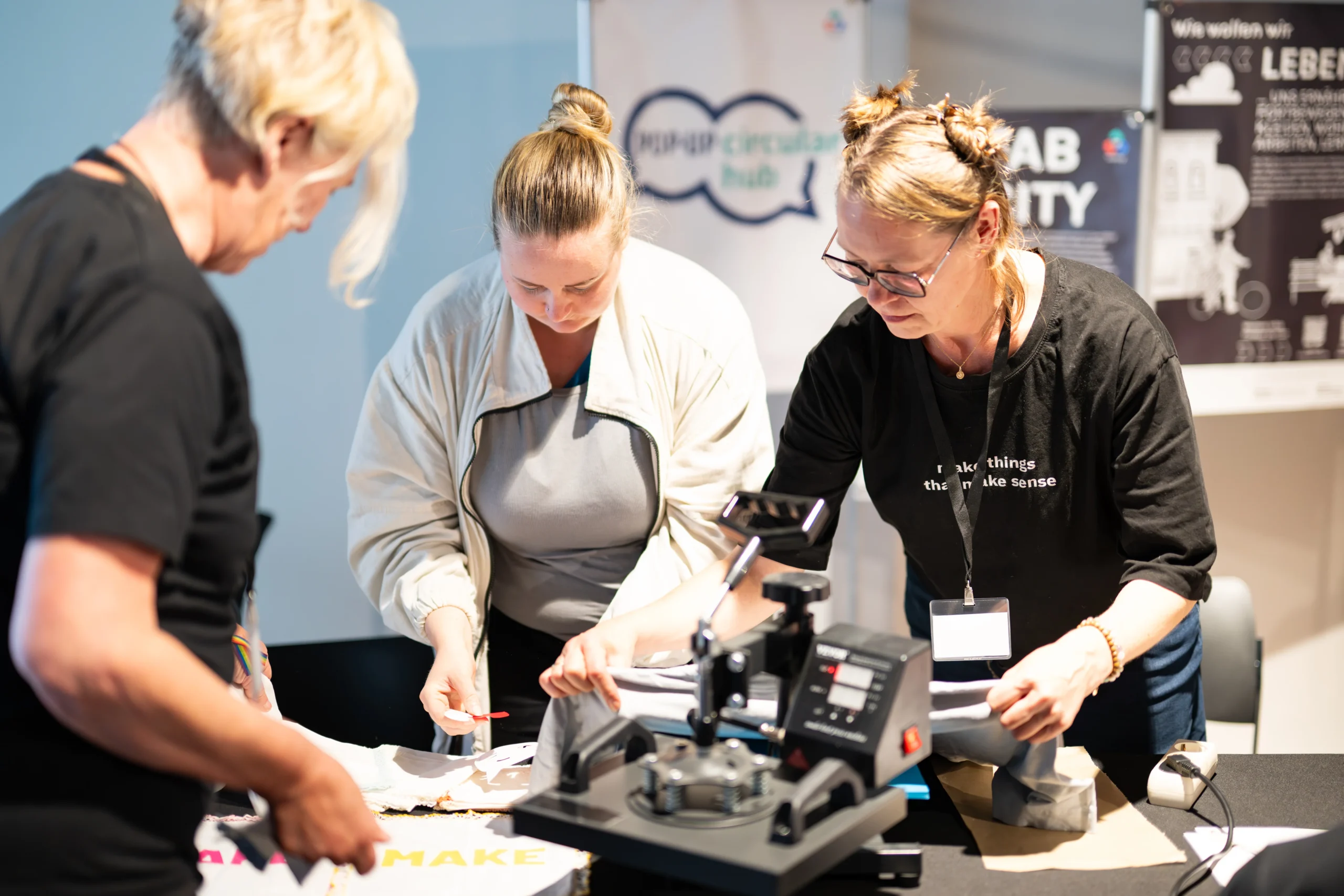 Drei Frauen arbeiten konzentriert an einem Tisch mit einer Maschine, möglicherweise bei einem Workshop oder Event von Fab City Hamburg.