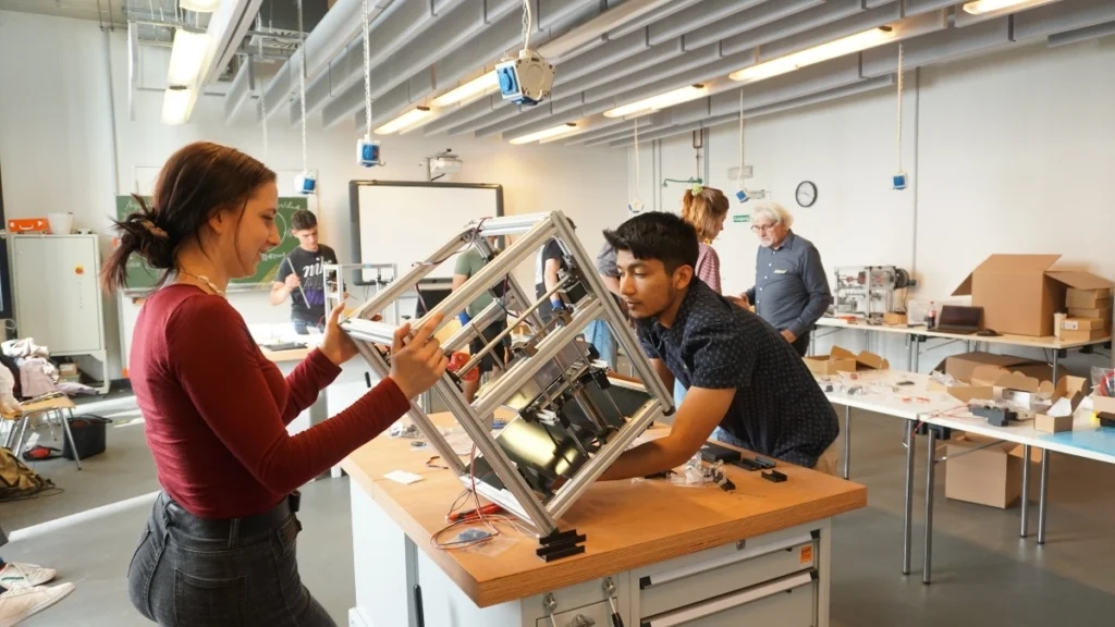 Zwei junge Menschen montieren gemeinsam eine Metallkonstruktion in einem hellen Werkstattraum, umgeben von weiteren Projektteilnehmern.