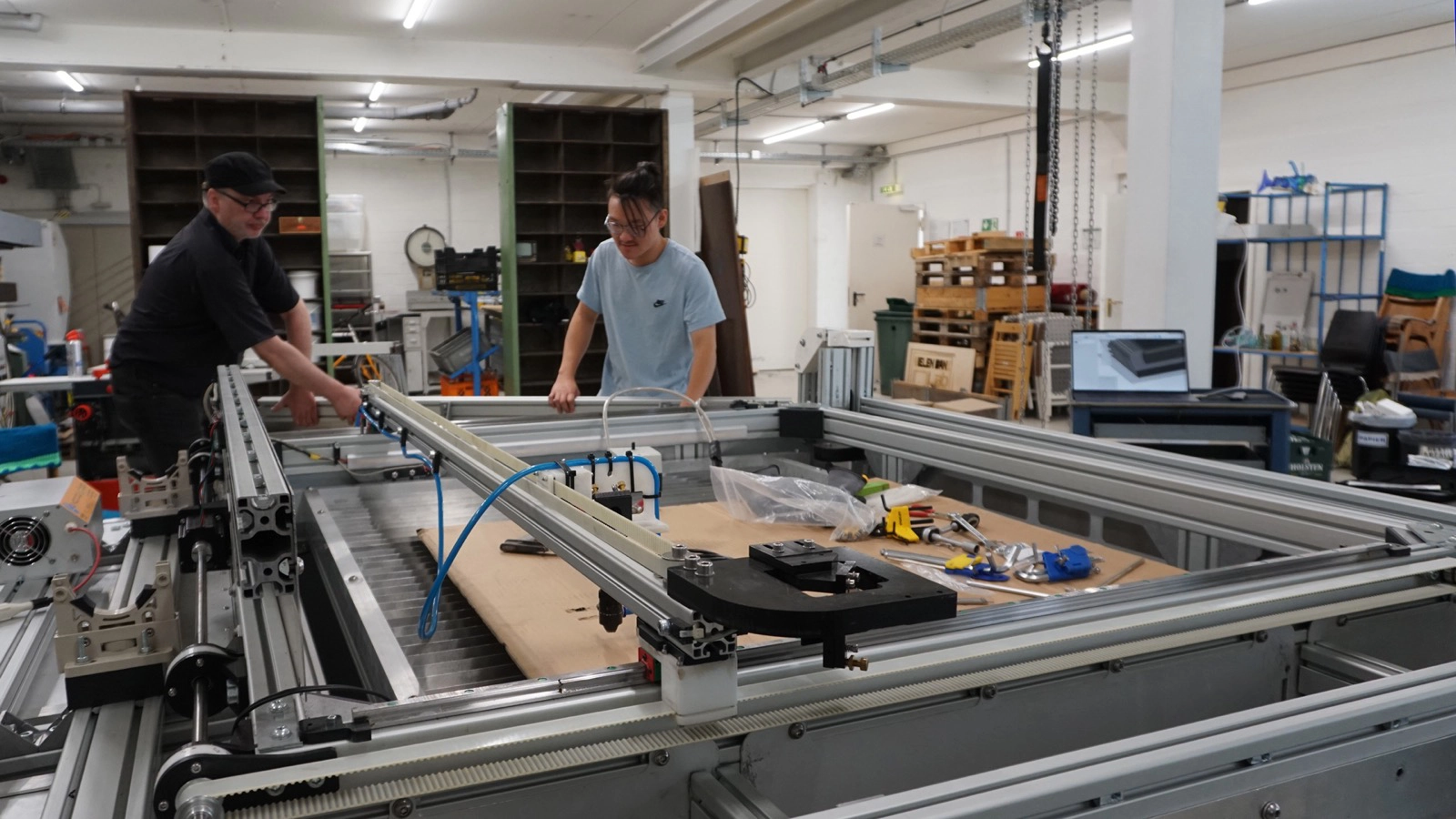 Zwei Männer von Fab City Hamburg arbeiten in einer Werkstatt an einer großen CNC-Maschine, während im Hintergrund ein Laptop ein 3D-Modell anzeigt.