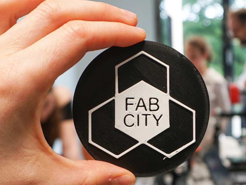 Eine Hand hält eine runde, schwarze Plakette mit dem weißen Schriftzug 'FAB CITY' und einem hexagonalen Muster.