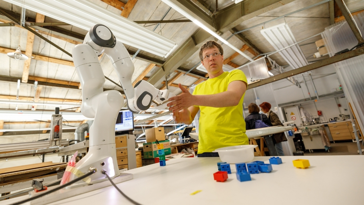 Ein Mann in einem gelben T-Shirt interagiert in einer Werkstatt der Fab City Hamburg mit einem weißen Roboterarm, umgeben von bunten Bausteinen.