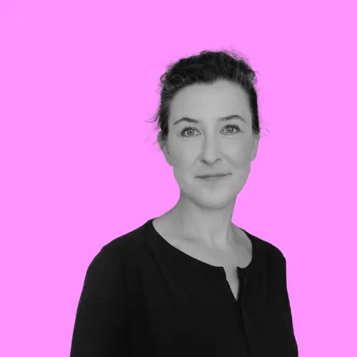 Porträt von Carolin Zastrow, einer Frau mit dunklen Haaren und einem leichten Lächeln, vor einem leuchtend pinken Hintergrund.