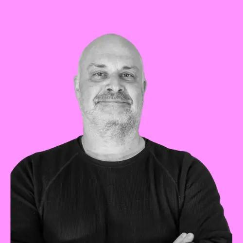 Porträt von Jan Buchenau, einem Mann mit Glatze und Bart, der vor einem pinkfarbenen Hintergrund lächelt und die Arme verschränkt hat.