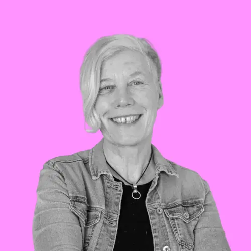 Porträt von Karen Loehnert, einer lächelnden Frau mit kurzen hellen Haaren und Jeansjacke, vor einem leuchtend pinken Hintergrund.