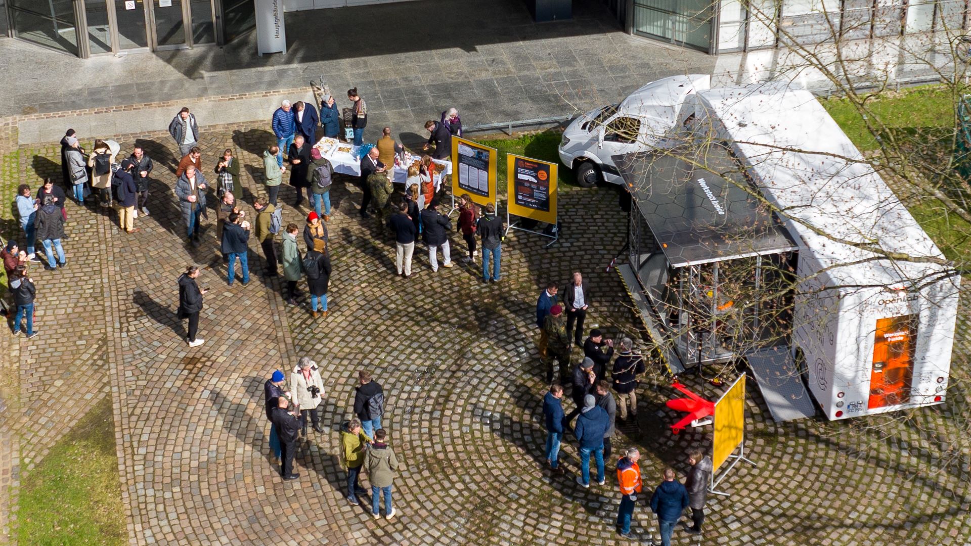 Viele Menschen versammeln sich auf einem gepflasterten Platz um ein mobiles Labor und Informationsstände bei einem Outdoor-Event.