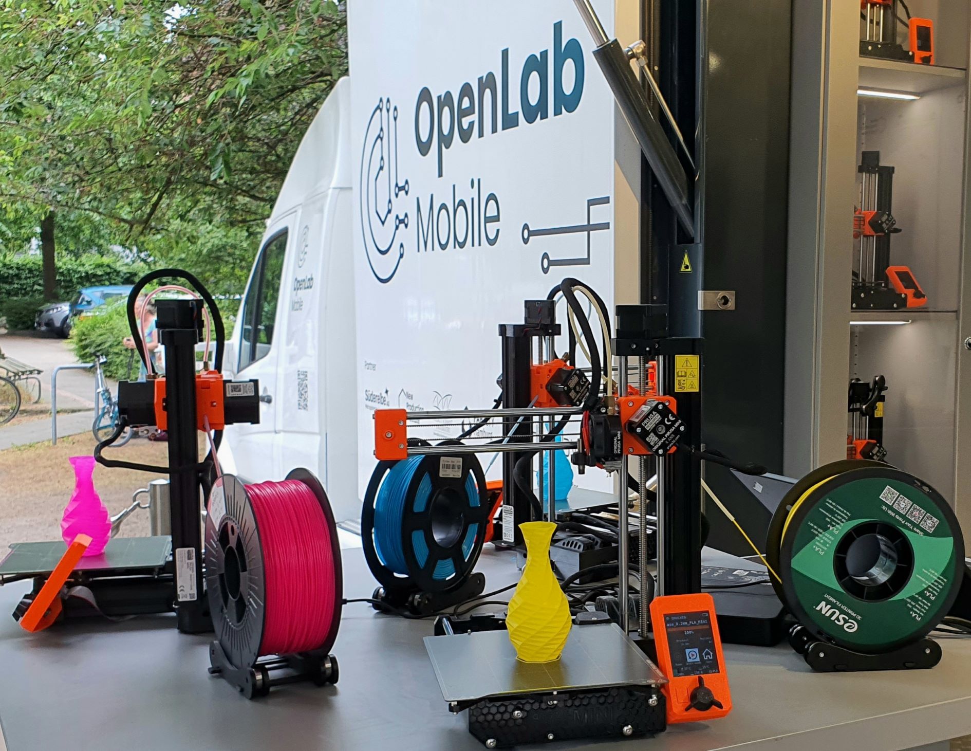 Drei 3D-Drucker mit farbigem Filament und gedruckten Objekten stehen auf einem Tisch vor einem weißen OpenLab Mobile Van im Freien.