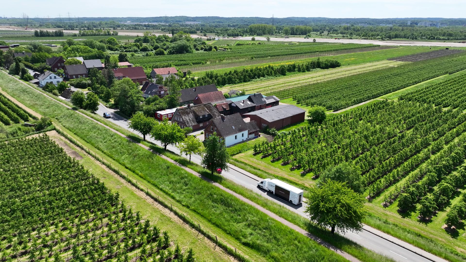 Luftaufnahme einer ländlichen Gegend mit Apfelplantagen, Wohnhäusern und einer Straße, auf der ein weißer Lieferwagen fährt.
