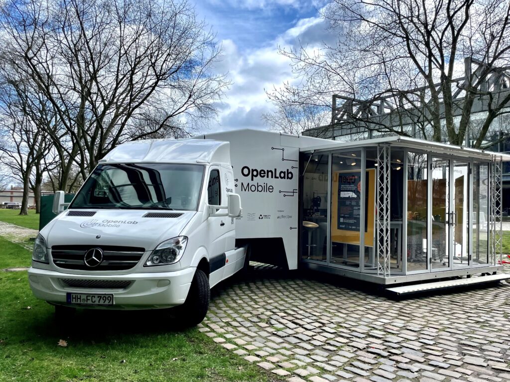 Ein weißer Mercedes-Benz Sprinter des OpenLab Mobile steht neben einer mobilen Glasausstellung von Fab City Hamburg auf einem gepflasterten Weg.