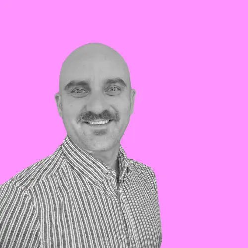 Porträt von Sascha Kargoscha, einem lächelnden Mann mit Glatze und Schnurrbart, vor einem pinkfarbenen Hintergrund.