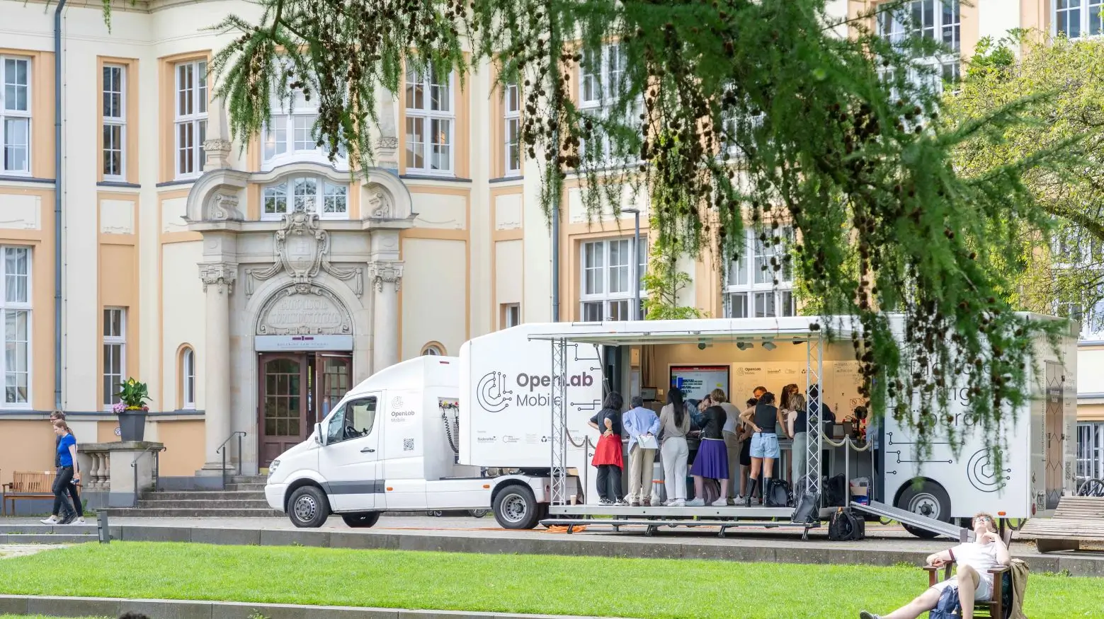 Ein weißer OpenLab Mobile Truck steht vor einem historischen Gebäude, während Menschen im Inneren interagieren und ein Ast ins Bild ragt.