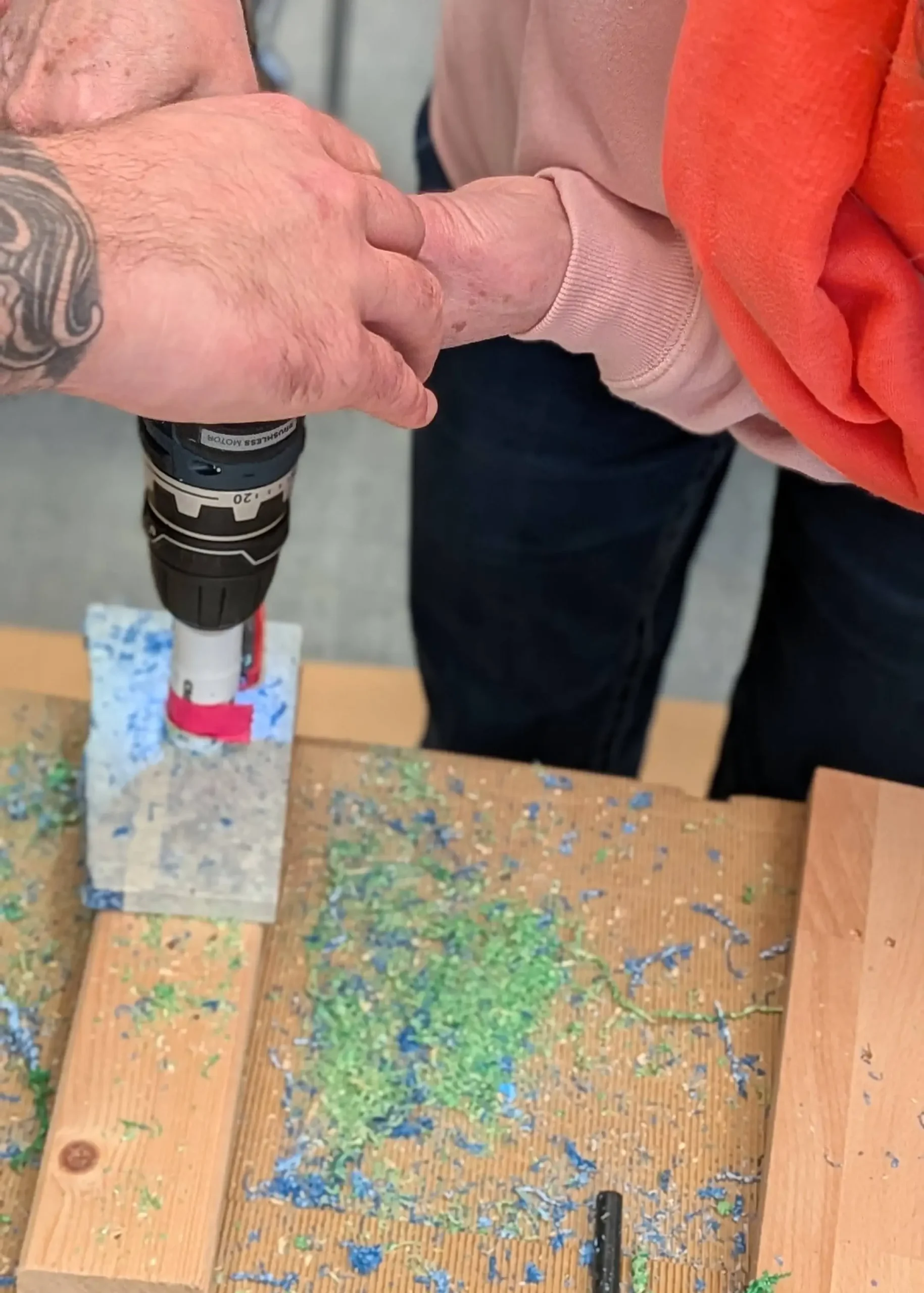 Zwei Hände bohren mit einem Akkuschrauber in einen Block aus recyceltem Material, wobei blaue und grüne Späne entstehen.