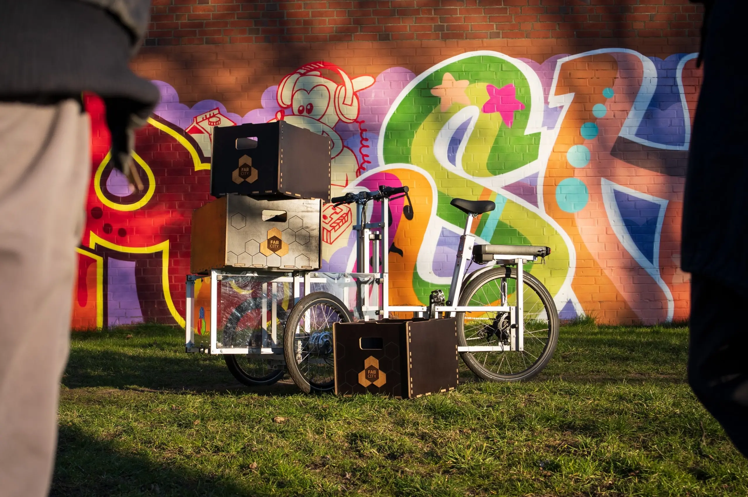 Ein Lastenrad von Fab City Hamburg mit Holzkisten steht auf Gras vor einer bunten Graffiti-Wand, teilweise von Personen verdeckt.