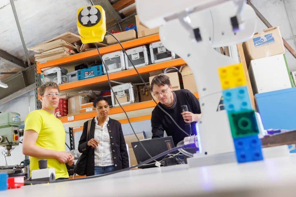 Drei Personen von Fab City Hamburg beobachten einen Roboterarm, der bunte Bausteine bewegt, in einem FabLab oder einer Werkstatt.