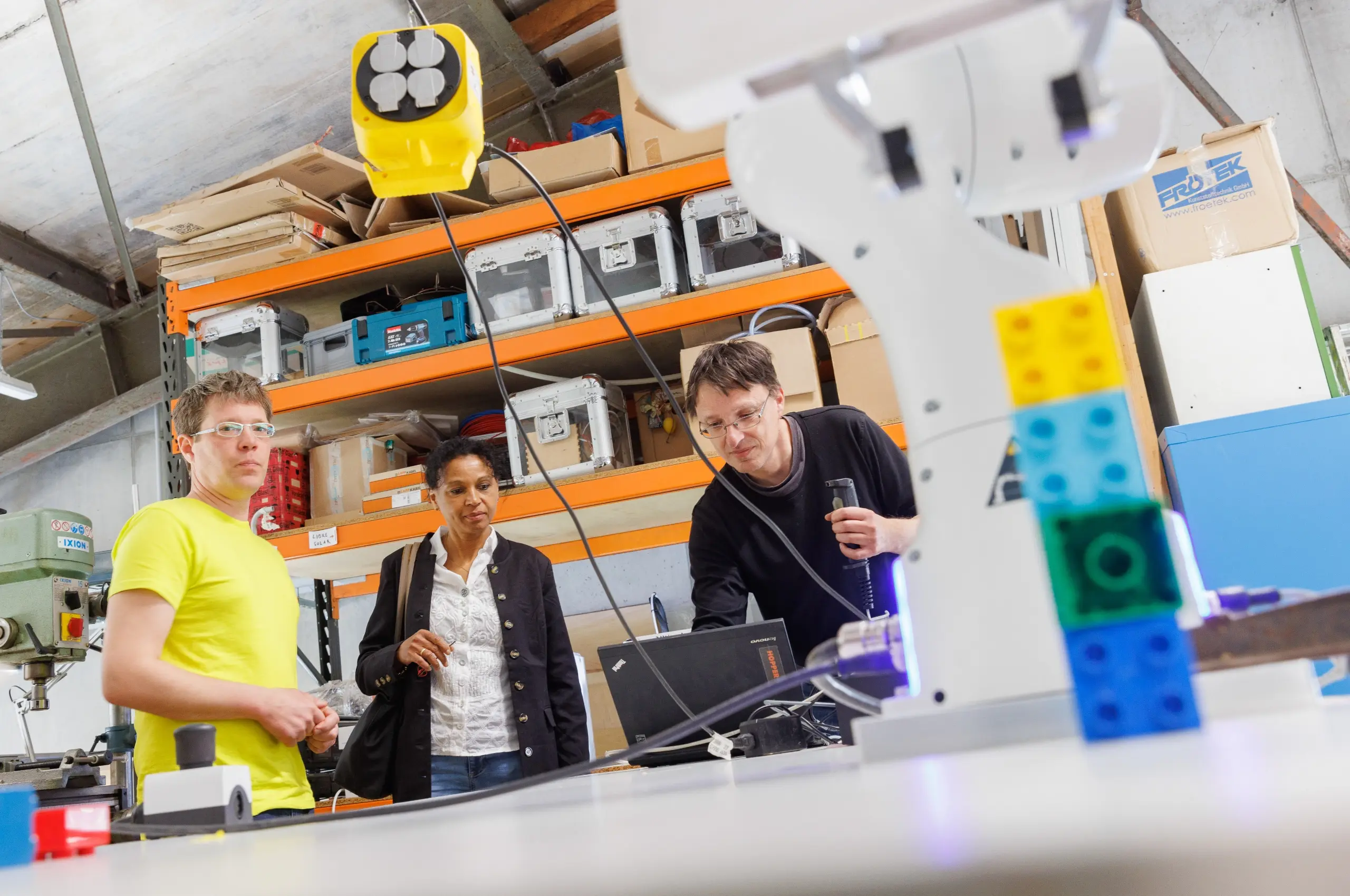 Drei Personen von Fab City Hamburg beobachten einen Roboterarm, der bunte Bausteine bewegt, in einem FabLab oder einer Werkstatt.