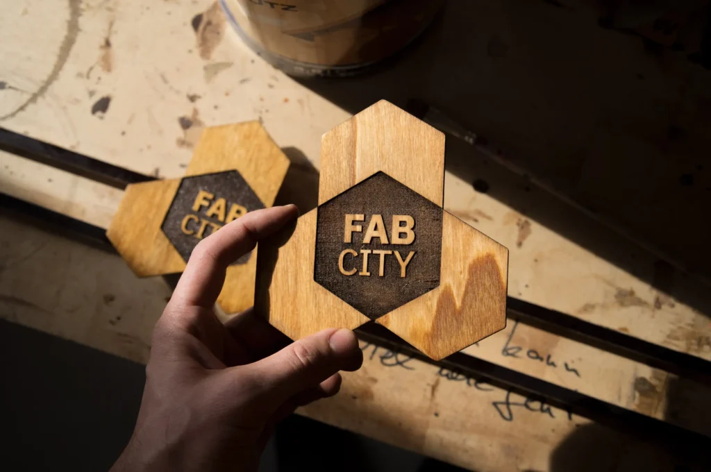 Eine Hand hält ein sechseckiges Holzlogo von Fab City Hamburg, das auf einer Werkbank liegt, mit einem weiteren Logo im Hintergrund.