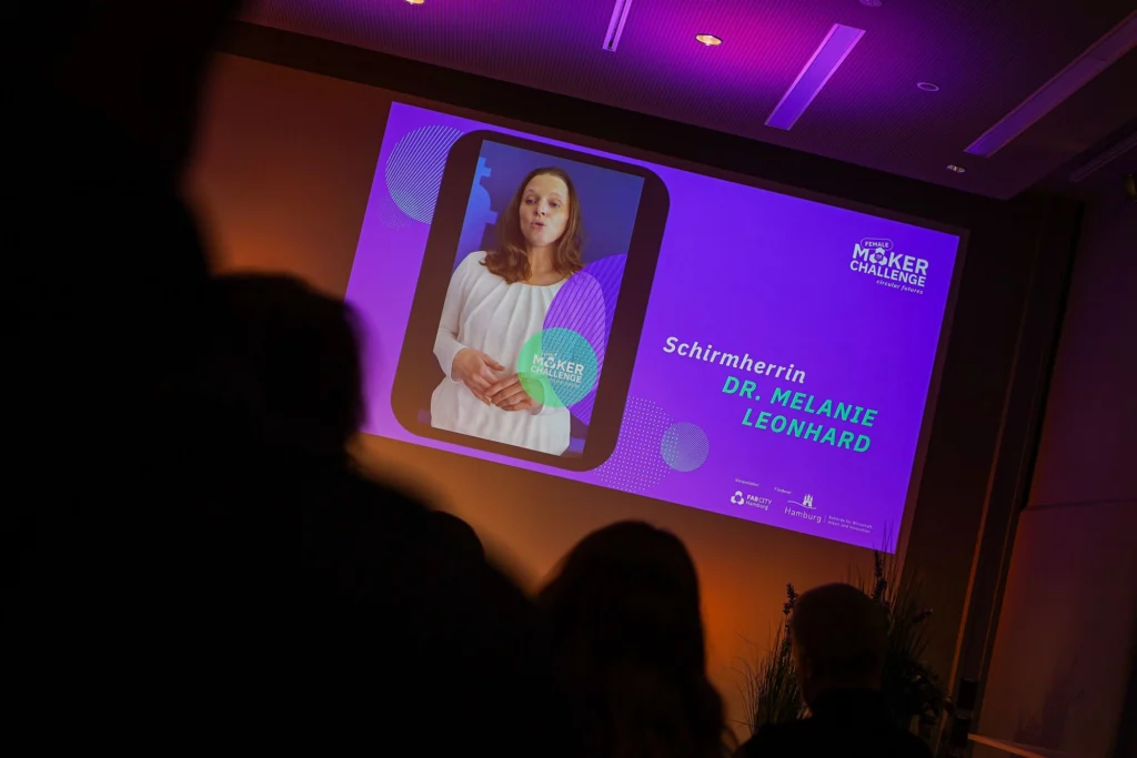 Ein Bildschirm zeigt Dr. Melanie Leonhard als Schirmherrin der Female Maker Challenge, während Silhouetten von Zuschauern im Vordergrund zu sehen sind.
