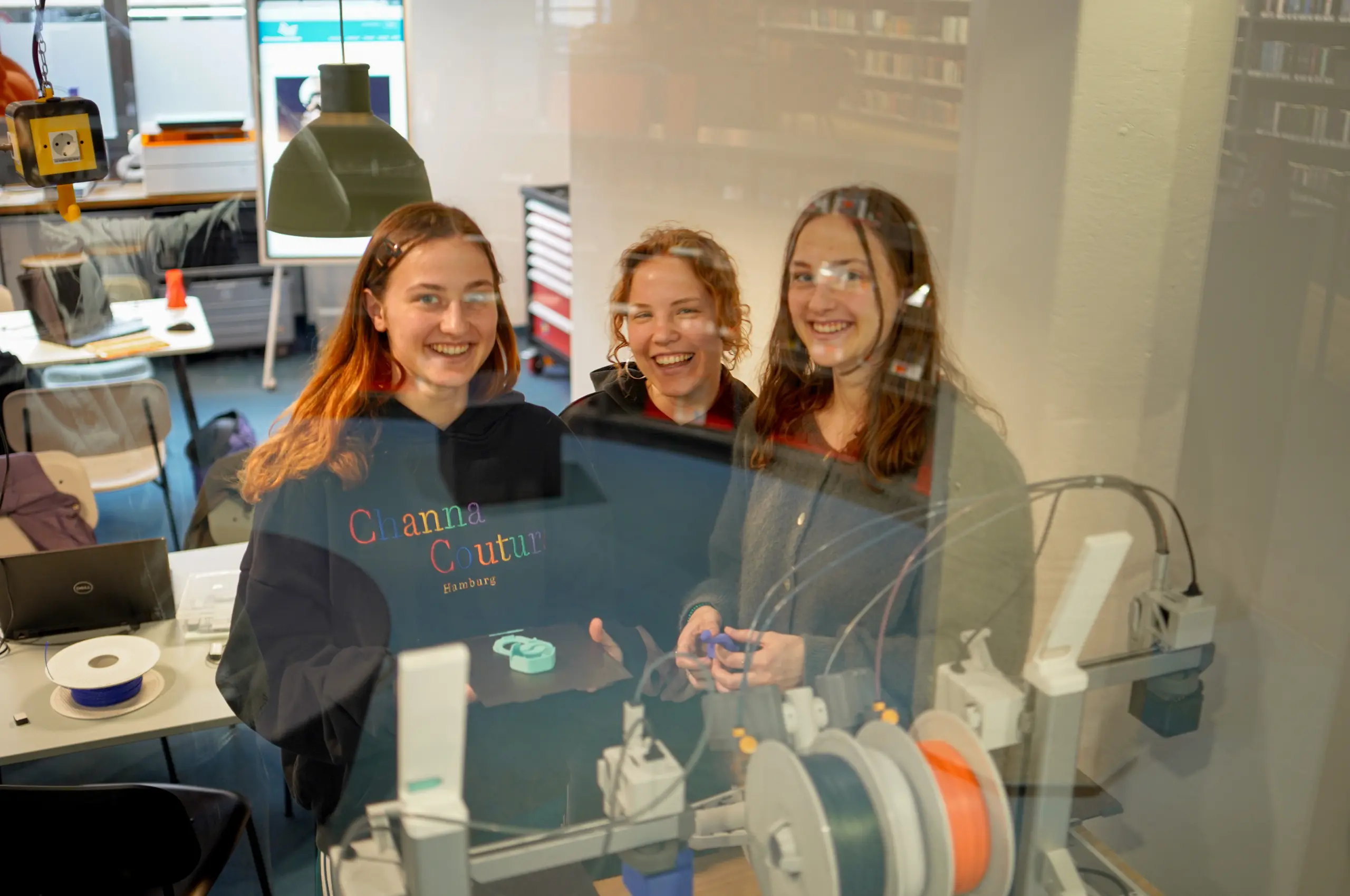 Drei junge Frauen lächeln im Fab City Hamburg Maker-Lab, umgeben von 3D-Druckern und Werkzeugen, während eines Female Maker Club Events.