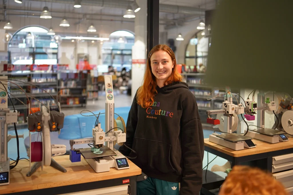 Eine lächelnde junge Frau mit rötlichen Haaren steht vor mehreren 3D-Druckern in einem FabLab der Bücherhalle Hamburg.