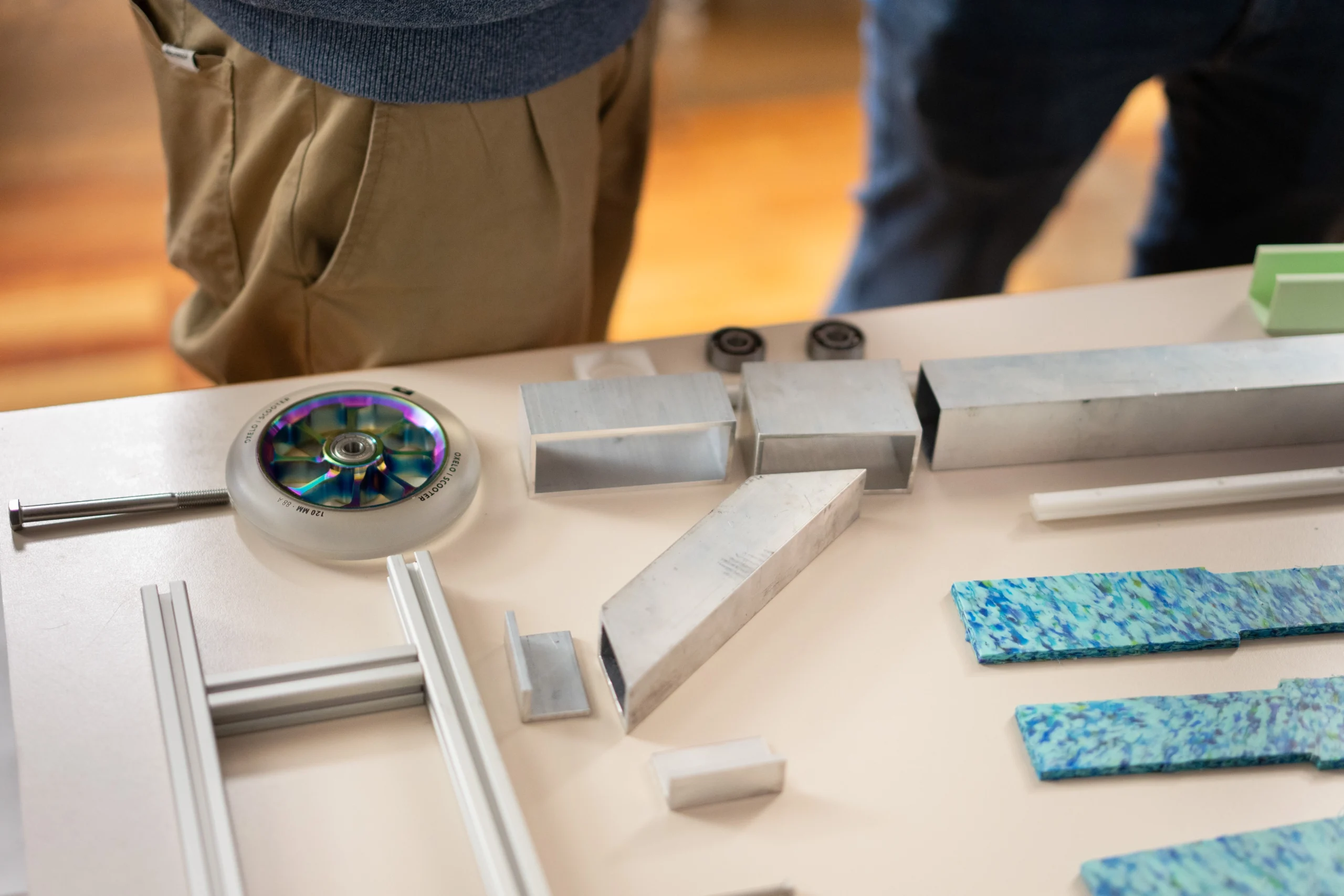 Verschiedene Bauteile wie ein Scooter-Rad, Aluminiumprofile und recycelte Kunststoffplatten liegen auf einem Tisch, umgeben von Personen bei einem Maker-Treffen