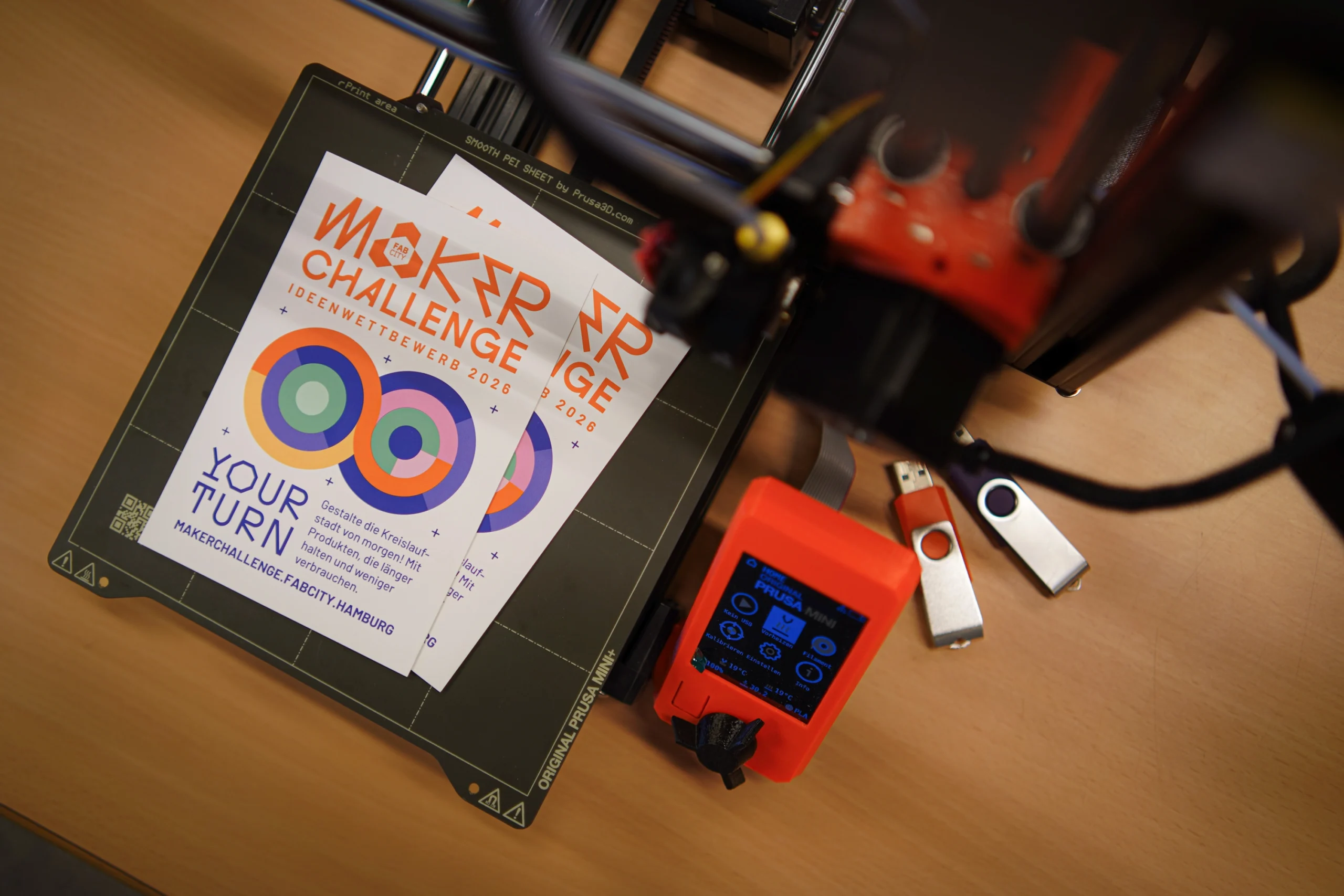 Flyer für die Fab City Maker Challenge 2026 liegen auf dem Druckbett eines 3D-Druckers, daneben ein Steuergerät und USB-Sticks.