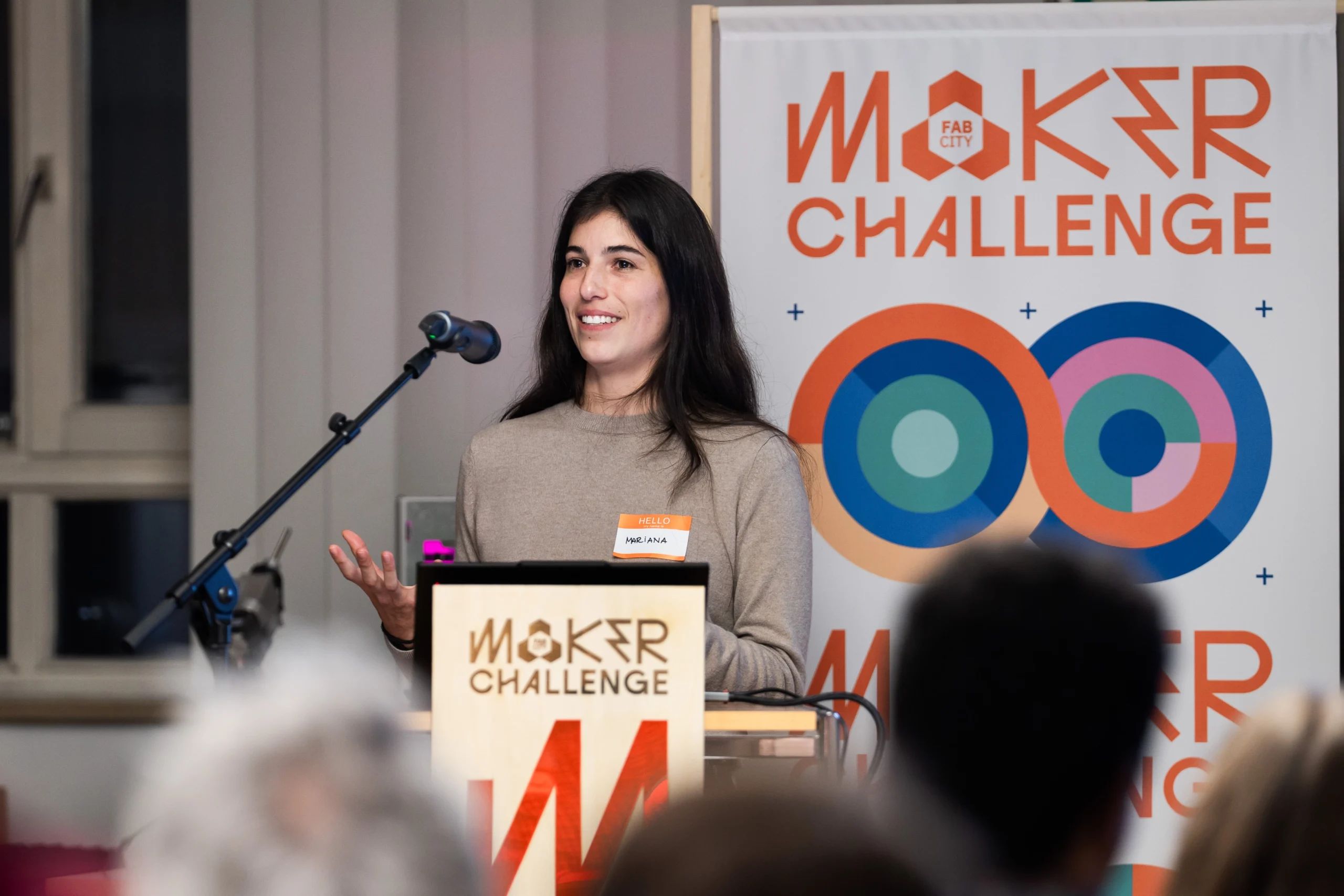 Mariana hält einen Vortrag bei der Maker Challenge, lächelnd vor einem Mikrofon und einem Banner mit dem Event-Logo.