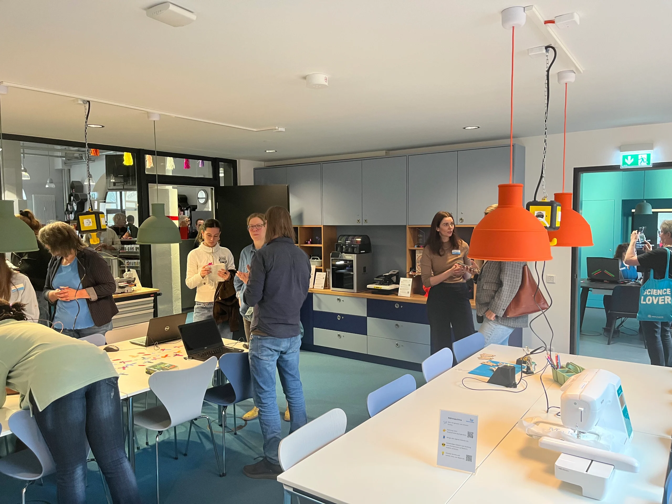 Menschen versammeln sich in einem modernen Maker Lab mit Tischen, Laptops, einer Nähmaschine und einem 3D-Drucker für kreative Aktivitäten.