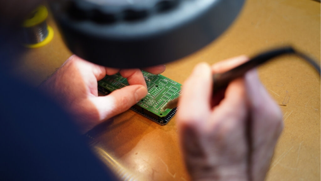 Eine Person lötet unter einer Lupe an einer grünen Leiterplatte, wobei die Hände und das Werkzeug im Fokus stehen.