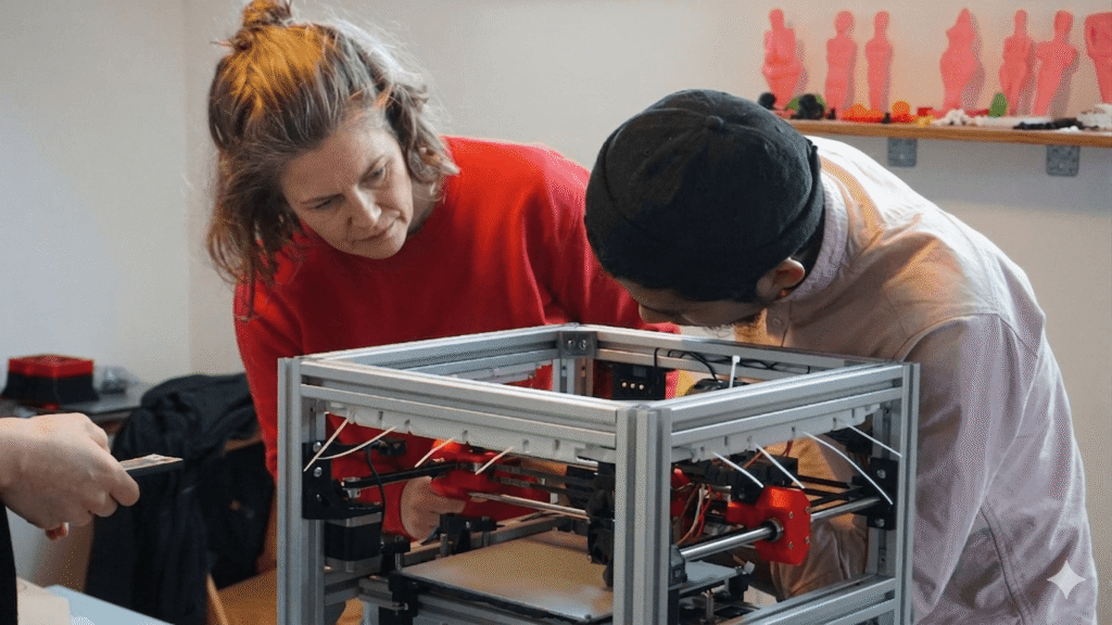 Zwei Personen beugen sich über einen 3D-Drucker und begutachten ihn, während im Hintergrund pinkfarbene Figuren auf einem Regal stehen.
