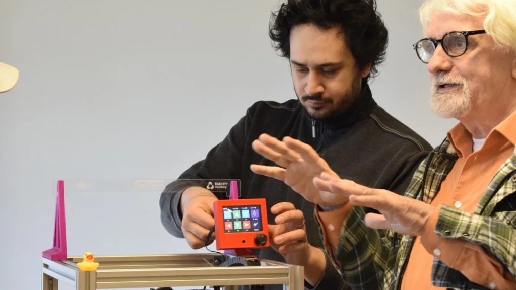 Ein jüngerer Mann bedient ein Steuergerät mit Fab City Hamburg Logo, während ein älterer Mann mit Brille und Bart gestikulierend spricht.